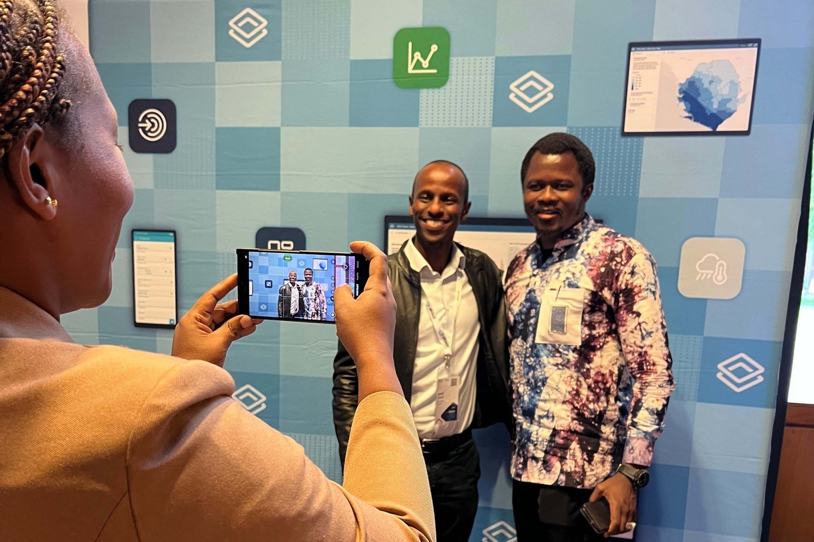A person takes a photo on their smartphone of two men posing in front of a blue event backdrop with white DHIS2 icons.