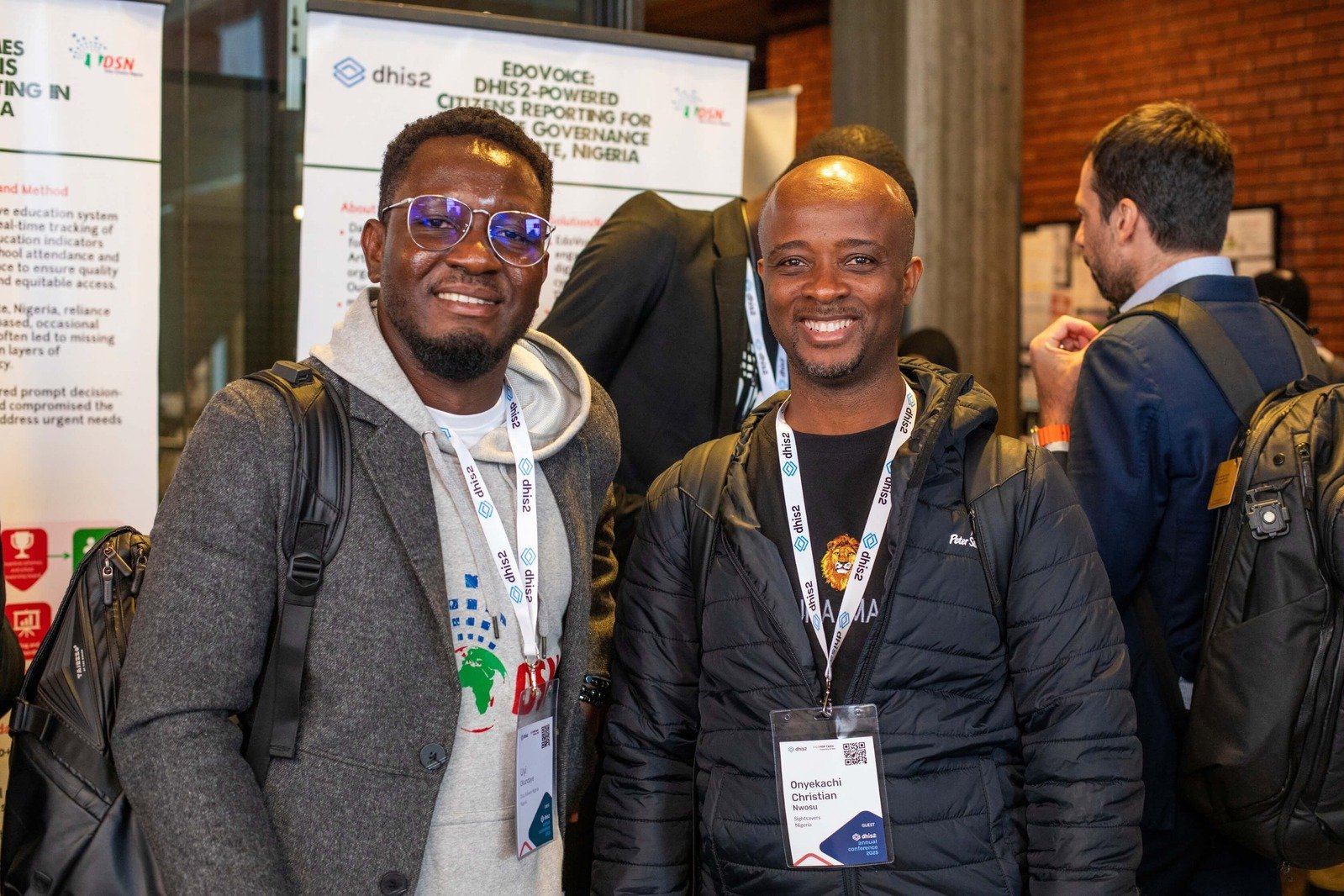 Two men, one wearing a grey hoodie and glasses, the other in a black jacket, smiling and posing with backpacks and conference lanyards at the DHIS2 Annual Conference.